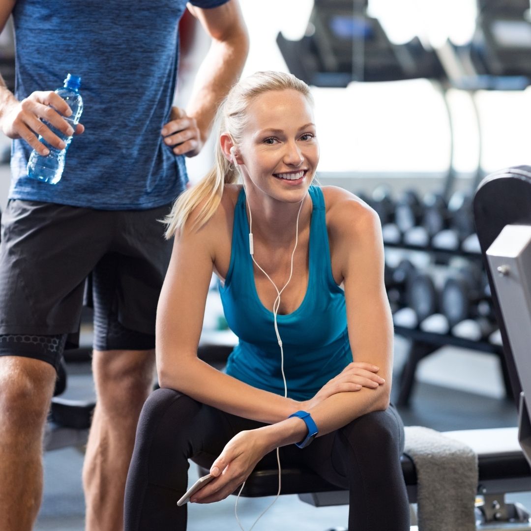 A woman with headphones smiles while resting on a gym bench after a workout.