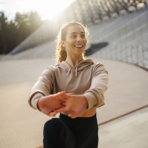 A smiling woman performs a dynamic arm and chest stretch outdoors during a sunny morning warm-up.